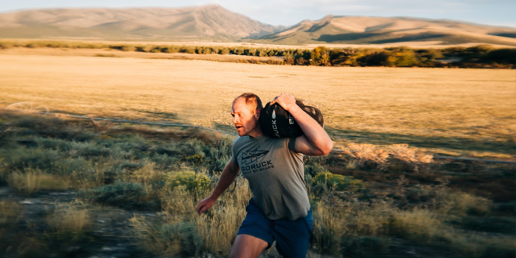 A man carries a Ruck up a hill with mountains in the background